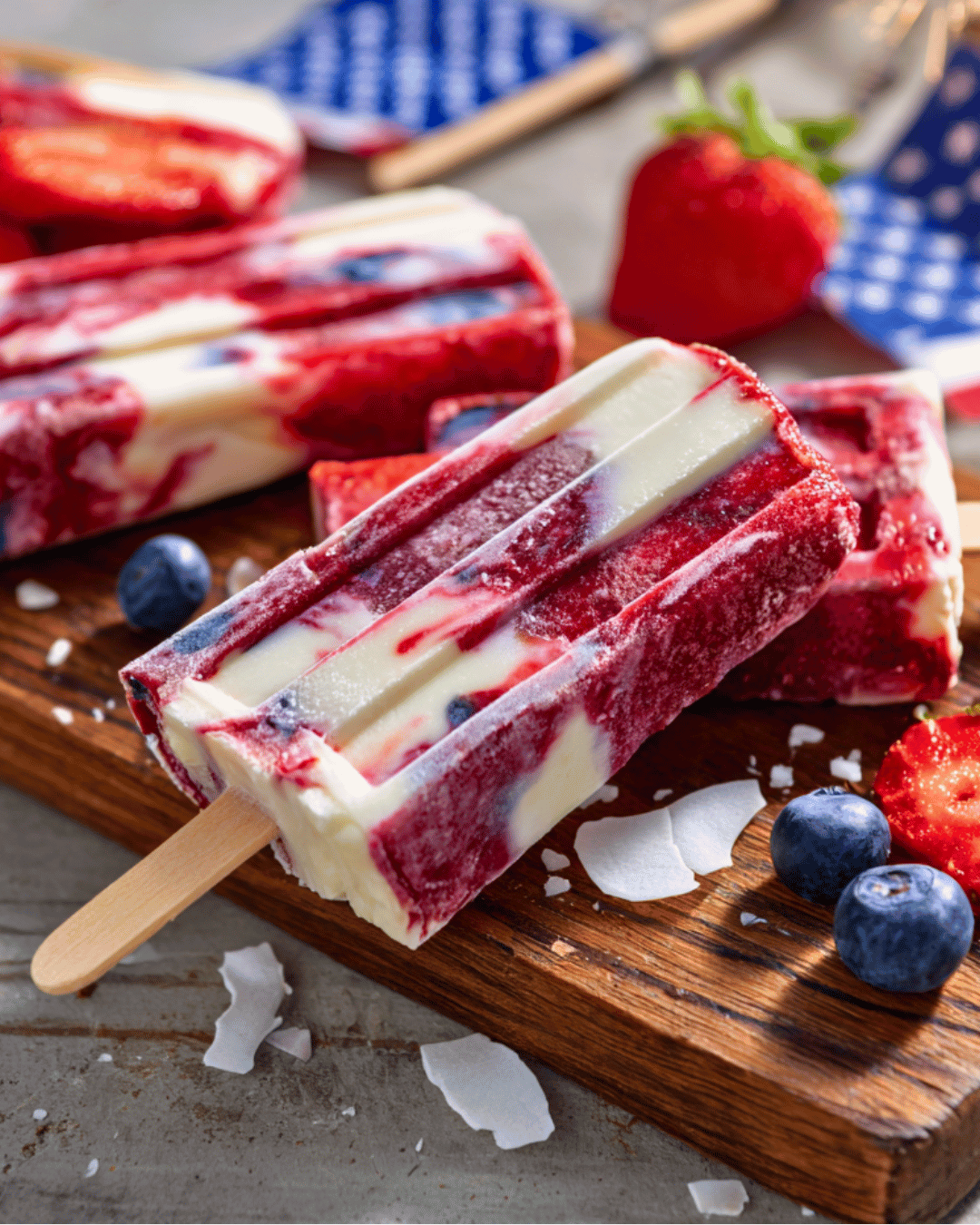 Red, white, and blue smoothie swirl popsicles with fruit in background on a wooden tray.